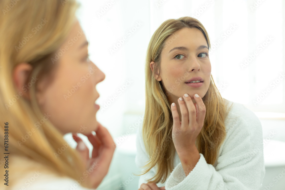 Happy caucasian woman in bathrobe looking in mirror and touching face in sunny bathroom Happy caucasian woman in bathrobe looking in mirror and touching face in sunny bathroom