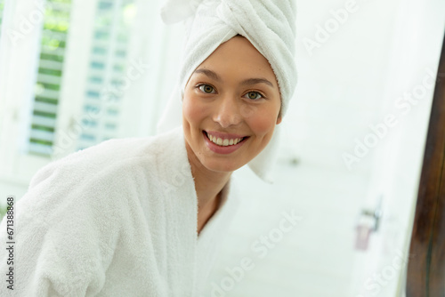Happy caucasian woman in bathrobe looking in mirror and smiling in sunny bathroom