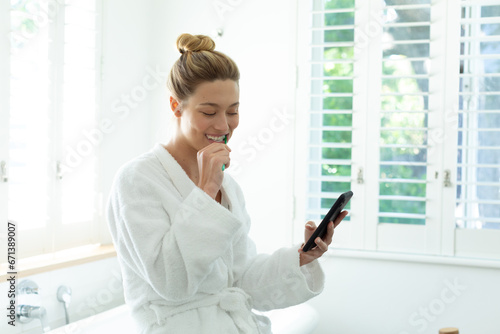 Happy caucasian woman in bathrobe brushing teeth and using smartphone in sunny bathroom