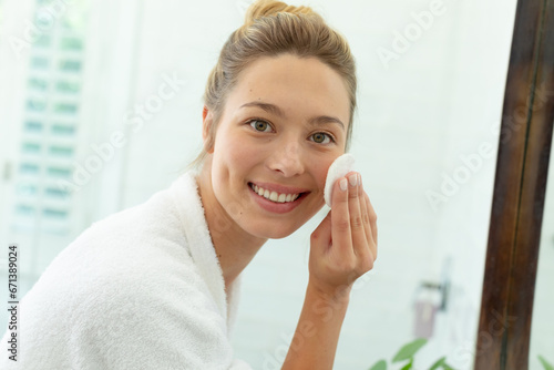Happy caucasian woman in bathrobe cleaning face with cotton pad and smiling in sunny bathroom