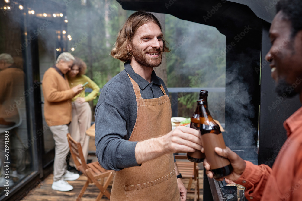 Happy young men drinking beer at party outdoors while they frying meat