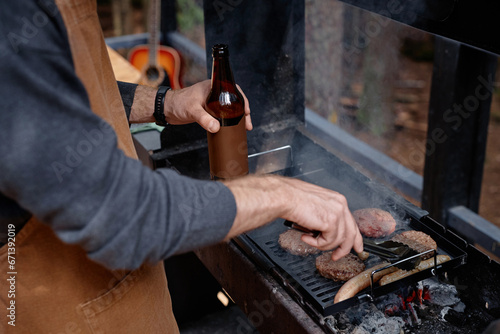 Close-up of young man drinking beer and frying meat on grill outdoors