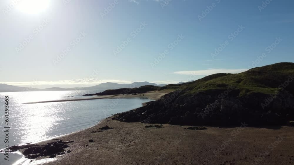 Aerial view rising right over Ynys Llanddwyn Welsh island with shimmering ocean and misty Snowdonia mountain range across the sunrise skyline Aerial view rising right over Ynys Llanddwyn Welsh island with shimmering ocean and misty Snowdonia mountain range across the sunrise skyline