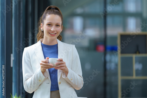 A businesswoman stand holding a cup of coffee and looking at a camera in an office. © Songsak C
