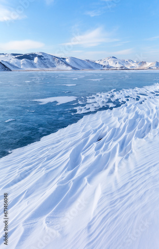 Scenic winter landscape of frozen Baikal Lake in February cold sunny day. Сoastal mountains are covered with snow. Snow crust in form of frozen waves. Natural cold background with empty space for text