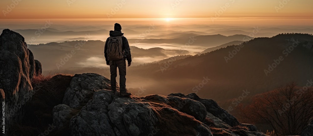 A mountaineer, standing alone on a rock at the top of a mountain enjoying the sunrise - obrazy, fototapety, plakaty 