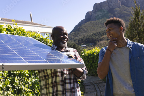 Happy african american father and adult son with solar panel talking in sunny garden photo