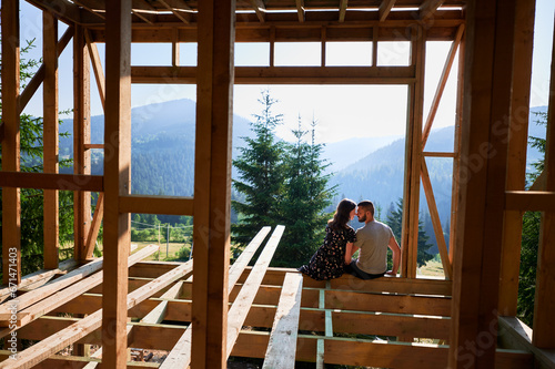 Man and woman inspecting their future wooden frame house nestled in the mountains near forest. Youthful couple at construction site in early morning. Concept of contemporary ecological construction.