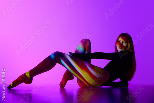 Sensual beautiful young girl in bodysuit posing against purple background with colorful neon lights reflection on body. Concept of modern art, beauty, style, futurism and cyberpunk, creativity
