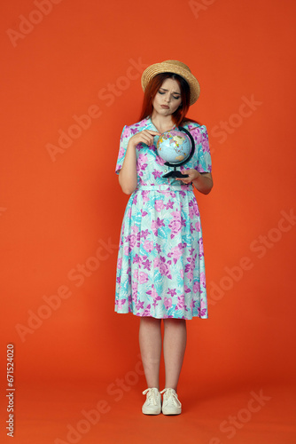 Woman holding small globe, sad face expression, pointing at the map picking destination, isolated on orange background. Portrait of female traveler.