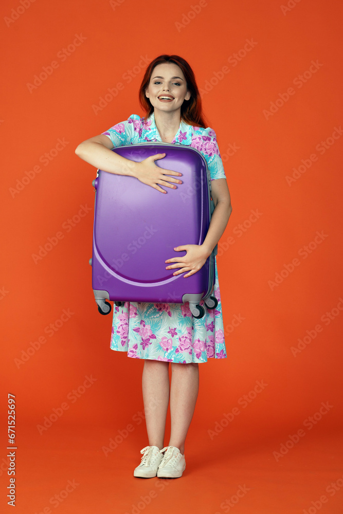 Woman standing holding big purple luggage case in hands, happy face smiling at the camera, isolated on orange background. Portrait of female traveler.