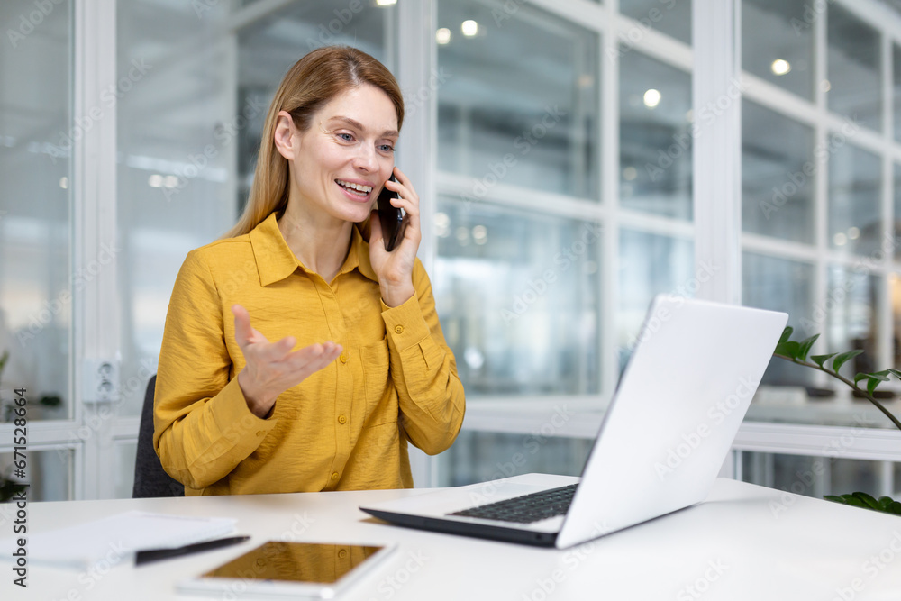 Mature successful blonde business woman in yellow shirt happily talking on the phone with a smile, female financier worker at the workplace in the middle of the office using a laptop at work. Mature successful blonde business woman in yellow shirt happily talking on the phone with a smile, female financier worker at the workplace in the middle of the office using a laptop at work.
