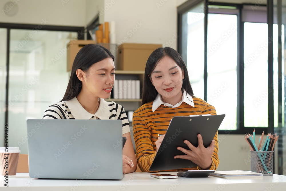 Two diverse mixed race beautiful african and asian business women wearing casual clothes, smiling with happiness, talking while taking break in indoor office, looking at laptop, holding cup of coffee Two diverse mixed race beautiful african and asian business women wearing casual clothes, smiling with happiness, talking while taking break in indoor office, looking at laptop, holding cup of coffee