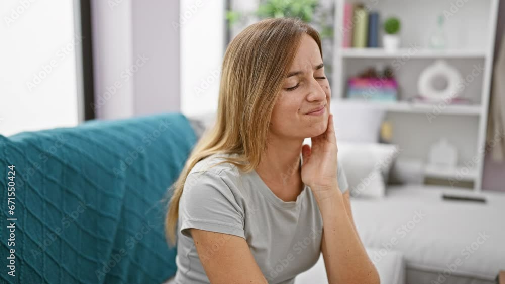 Attractive young blonde adult woman sitting on living room sofa at home, suffering in agony from severe teeth pain - oral issue demanding dentist's help urgently. Attractive young blonde adult woman sitting on living room sofa at home, suffering in agony from severe teeth pain - oral issue demanding dentist's help urgently.