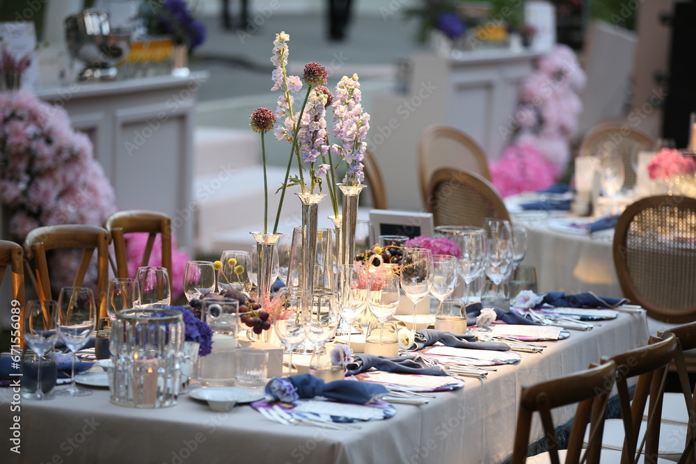Decorated white wedding table for a festive dinner with pink flowers in brass pots on green lawn under the open sky.