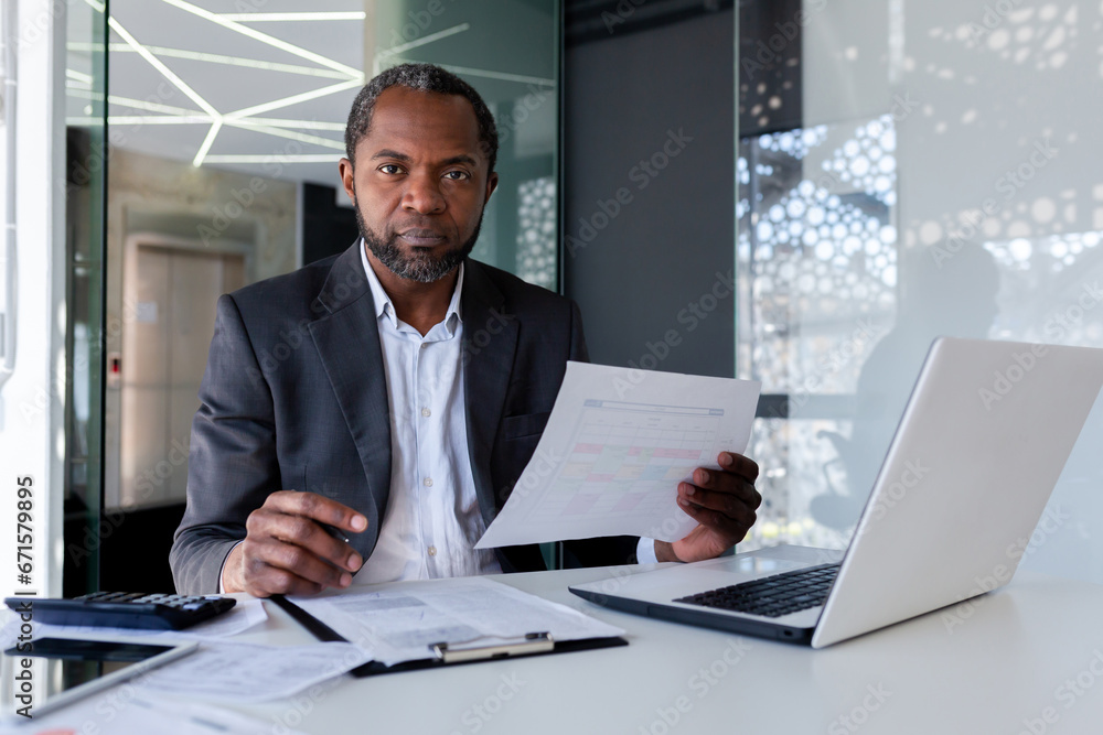 Portrait of mature strict serious financier businessman, man looking at camera with concentration and serious look at camera, boss in business suit behind paper work using laptop inside office - obrazy, fototapety, plakaty 