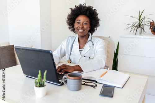 Attractive female doctor smiling looking at camera while working with laptop in the consultation.