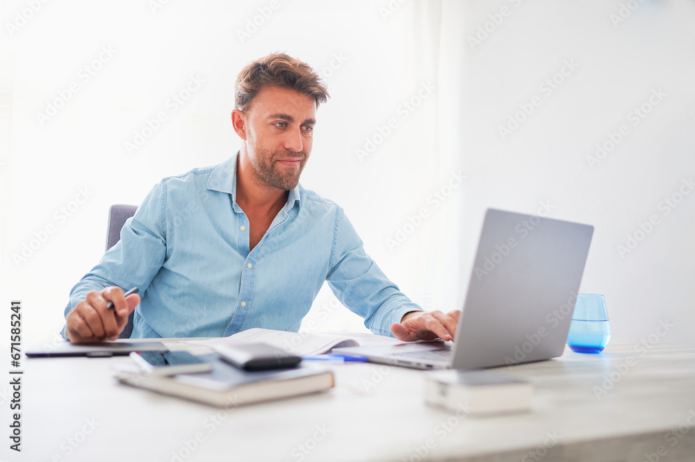 Smiling young confident man dressed in blue, using a laptop at his workplace in the office. Smiling young confident man dressed in blue, using a laptop at his workplace in the office.