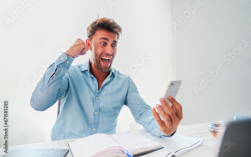 Excited young man dressed in shirt looks at smart phone and makes fist while celebrating winning a sports bet.