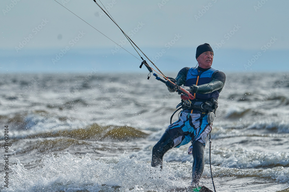 A mature male surfer rides a board with a kite on a windy autumn day across a large body of water. Splashes of water scatter to the sides. A mature male surfer rides a board with a kite on a windy autumn day across a large body of water. Splashes of water scatter to the sides.