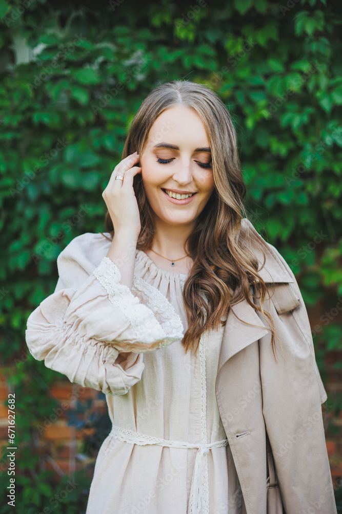 Portrait of a beautiful young woman in a light beige dress. Romantic woman in a dress with a smile on her face on the background of a green garden. Women's cocktail dress for a holiday Portrait of a beautiful young woman in a light beige dress. Romantic woman in a dress with a smile on her face on the background of a green garden. Women's cocktail dress for a holiday
