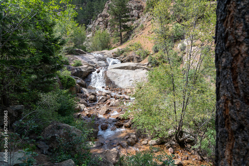 A waterfall cascade on the Seven Bridges Trail in North Cheyenne Cañon Park in Colorado Springs, CO in the late afternoon on a sunny summer day, with trees, red rocks, and mountains in the landscape
