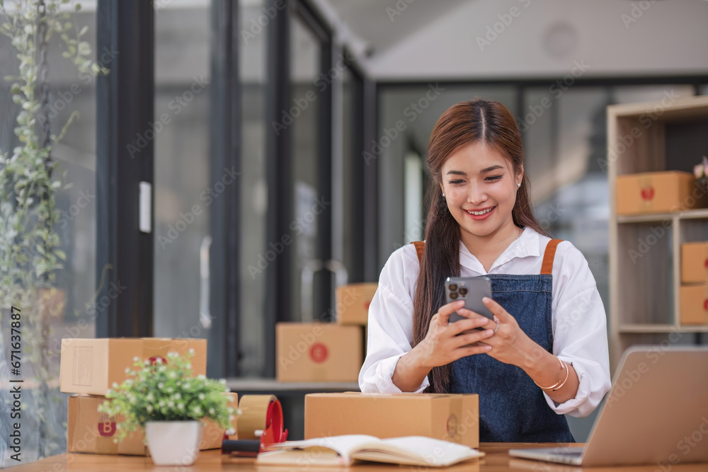 A portrait of a young Asian woman, e-commerce employee sitting in the office full of packages in the background write note of orders and use smartphone, for SME business ecommerce and delivery A portrait of a young Asian woman, e-commerce employee sitting in the office full of packages in the background write note of orders and use smartphone, for SME business ecommerce and delivery