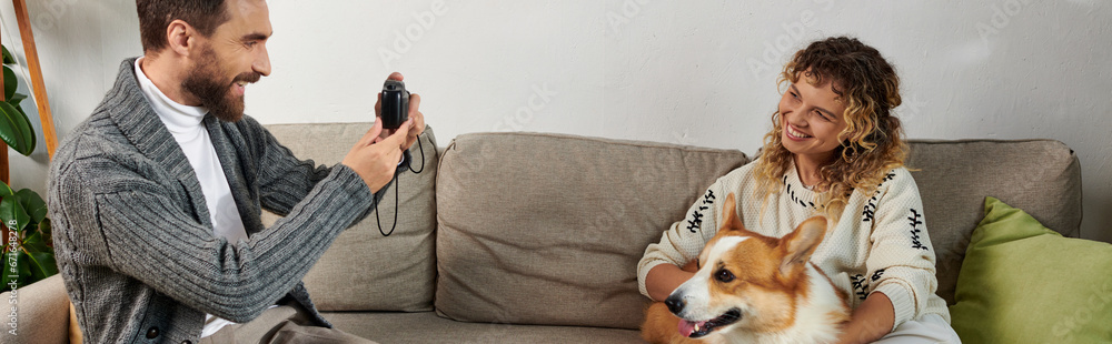 happy man smiling and taking photo of wife with corgi dog on camera in modern apartment, banner happy man smiling and taking photo of wife with corgi dog on camera in modern apartment, banner