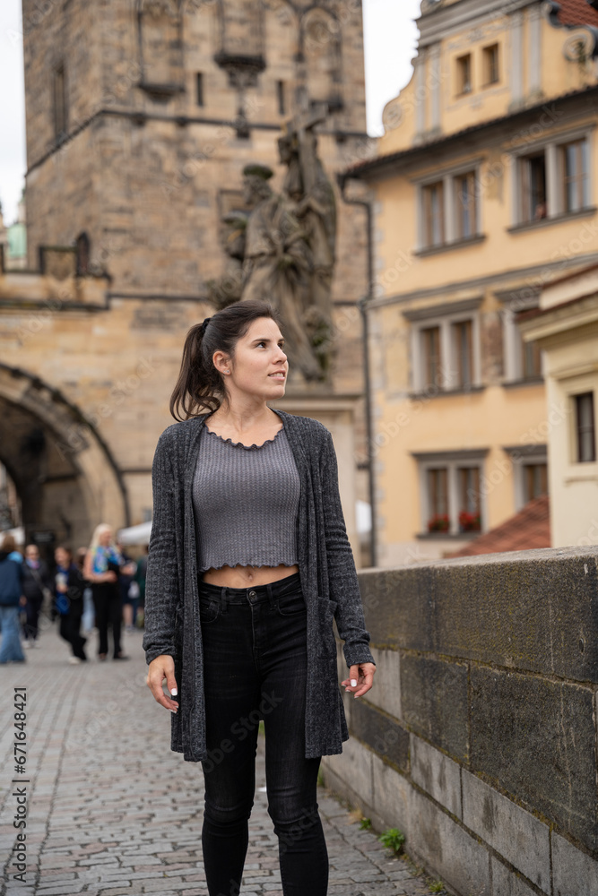 Fototapeta premium Young female tourist walking on the Charles Bridge in Prague, wearing dark clothes on a cloudy day.