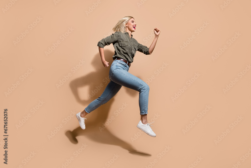 Full length photo of impressed funky woman dressed khaki shirt running fast jumping high isolated beige color background