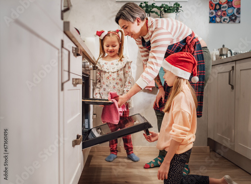 Cute little girls in red Santa hats with mother making homemade sweet biscuits. They putting baking sheet in hot oven. Christmas cookies preparation. Home sweet home warm family life moments concept..