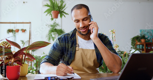 African american male worker of flower shop sitting at table and calling on smartphone and writing down in planner while sitting in cozy floral shop. Caucasian young male florist flower store owner