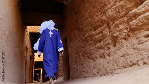 A traditionally dressed Bedouin Moroccan man wearing a blue gandoura and a turban walks inside the Kasbah in Mhmaid, Morocco. Authentic rural scene of South Morocco. 4k.   photo