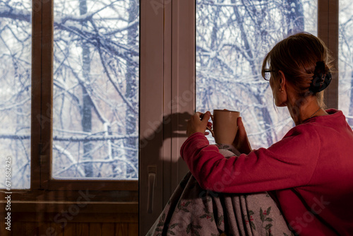 woman drinks coffee on an early winter morning and admires snow-covered trees outside her window. Hygge, cozy photo