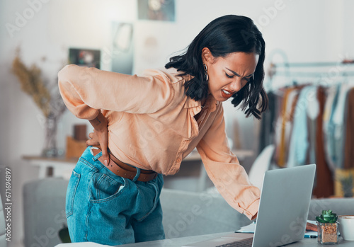 Back pain, discomfort and tired designer working on a laptop in a clothing boutique or store. Young overworked business owner, shop assistant suffering from injury, backache and pain at desk photo