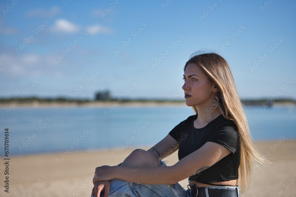 Portrait of young, beautiful blonde woman, green eyes, with black top, torn jeans and tattoos, sitting, looking at infinity with the sea in the background. Concept of peace, tranquility, relaxation.