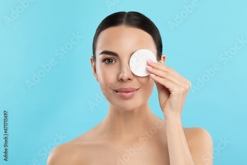 Beautiful woman removing makeup with cotton pad on light blue background