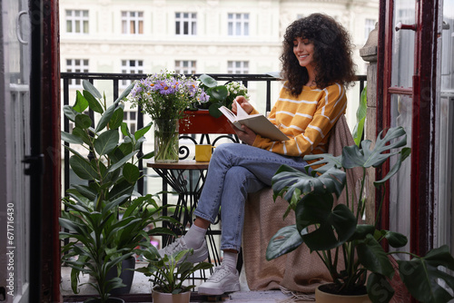 Young woman reading book at table on balcony with beautiful houseplants