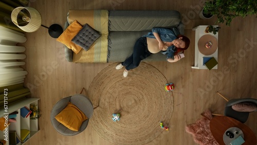 In the shot from above a pregnant woman lies down on a sofa in a room. She holds the phone, looks at something in it and smiles. She is happy and resting. There are childrens toys on the floor nearby