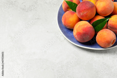 Many whole fresh ripe peaches and green leaves in plate on white table, space for text