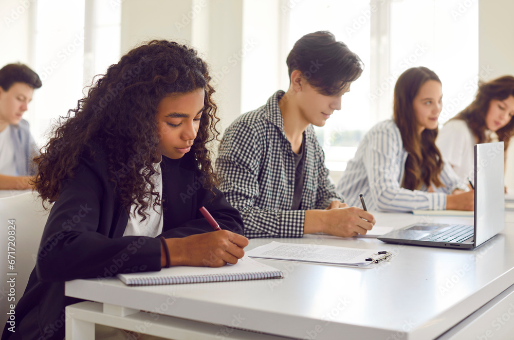 Side view of a group of diverse young high school students sitting at the desk in a row and writing notes during a college lesson in the classroom. Education and knowledge concept. - obrazy, fototapety, plakaty 