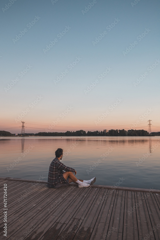 Young 25 year old brown man with plaid shirt sitting on the end of a wooden pier during sunset, watching the calm water surface in Almere Netherlands