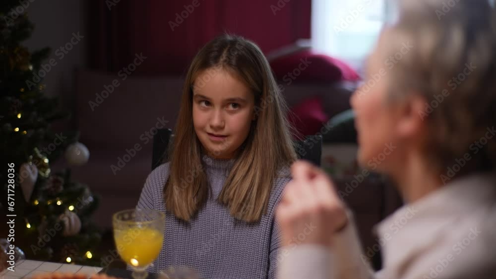 Adorable little girl sitting on Christmas family party, eating, chatting and smiling at dinner holiday table in room with cozy interior on xmas eve, on background of decorating tree with lights. Adorable little girl sitting on Christmas family party, eating, chatting and smiling at dinner holiday table in room with cozy interior on xmas eve, on background of decorating tree with lights.