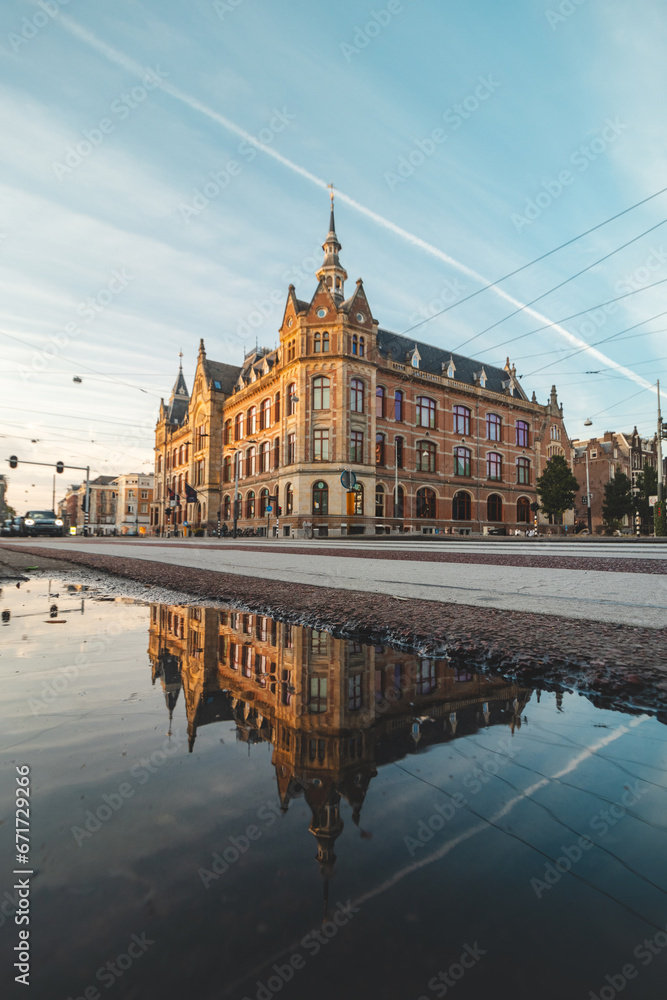 Fototapeta premium Historic building in the centre of Amsterdam under the light from the sunset at a busy intersection in the middle of the city with a reflection in a puddle