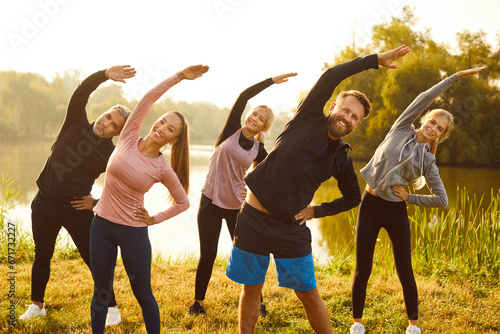 Group of people having an outdoor fitness workout. Five happy men and women in activewear doing sports exercises in nature. Smiling male and female athletes standing on a river bank doing a side bend