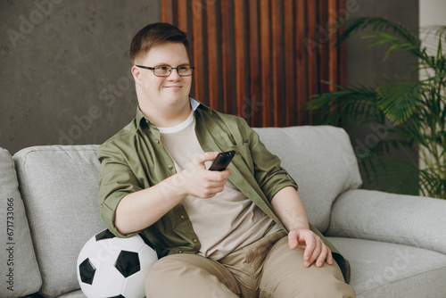 Young man with down syndrome wear glasses casual clothes watch tv football live stream sits on grey sofa couch stay at home flat rest spend free time in living room. Genetic disease world day concept.