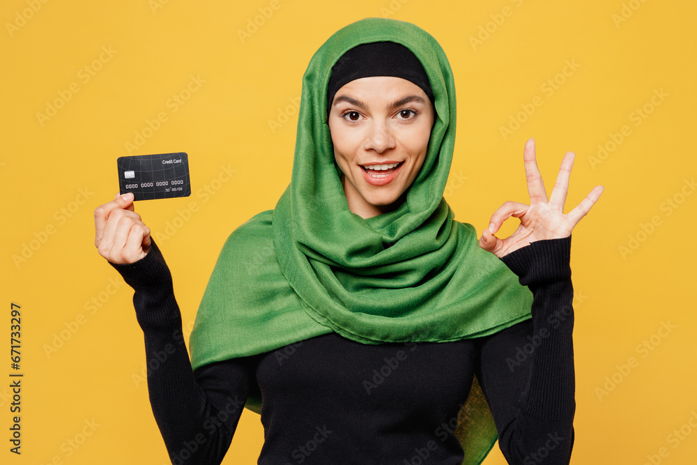 Young fun arabian asian muslim woman wearing green hijab abaya black clothes hold credit bank card show ok okay isolated on plain yellow background. People uae middle eastern islam religious concept. Young fun arabian asian muslim woman wearing green hijab abaya black clothes hold credit bank card show ok okay isolated on plain yellow background. People uae middle eastern islam religious concept.