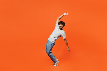Full body side view young smiling happy Indian man he wears t-shirt casual clothes stand on toes leaning back with outstretched hands isolated on orange red color background studio. Lifestyle concept.