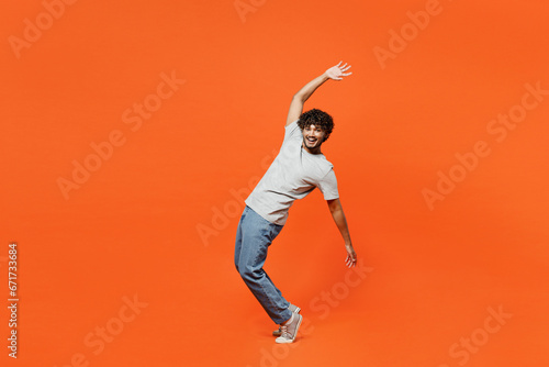 Full body side view young smiling happy Indian man he wears t-shirt casual clothes stand on toes leaning back with outstretched hands isolated on orange red color background studio. Lifestyle concept.