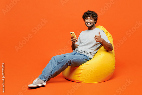 Full body young smiling fun happy Indian man he wears t-shirt casual clothes sit in bag chair use mobile cell phone type message show thumb up isolated on orange red color background studio portrait. photo